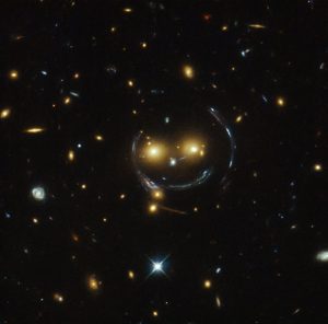In the centre of this image, taken with the NASA/ESA Hubble Space Telescope, are two faint galaxies that seem to be smiling. You can make out two orange eyes and a white button nose. In the case of this “happy face”, the two eyes are the galaxies SDSSCGB 8842.3 and SDSSCGB 8842.4 and the misleading smile lines are actually arcs caused by an effect known as strong gravitational lensing.  Massive structures in the Universe exert such a powerful gravitational pull that they can warp the spacetime around them and act as cosmic lenses which can magnify, distort and bend the light behind them. This phenomenon, crucial to many of Hubble’s discoveries, can be explained by Einstein’s theory of general relativity. In this special case of gravitational lensing, a ring  — known as an Einstein Ring  — is produced from this bending of light, a consequence of the exact and symmetrical alignment of the source, lens and observer and resulting in the ring-like structure we see here. Hubble has provided astronomers with the tools to probe these massive galaxies and model their lensing effects, allowing us to peer further into the early Universe than ever before. This object was studied by Hubble’s Wide Field and Planetary Camera 2 (WFPC2) and Wide Field Camera 3 (WFC3) as part of a survey of strong lenses. A version of this image was entered into the Hubble’s Hidden Treasures image processing competition by contestant Judy Schmidt.